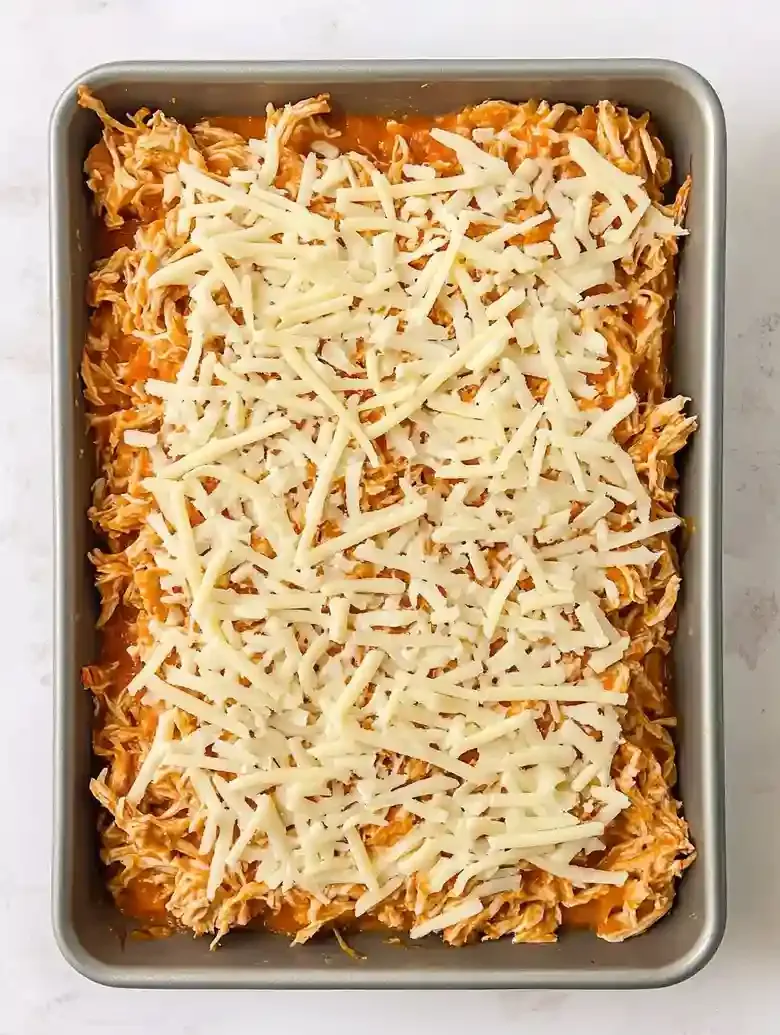 Baking tray filled with shredded chicken in orange sauce, topped with cheese.