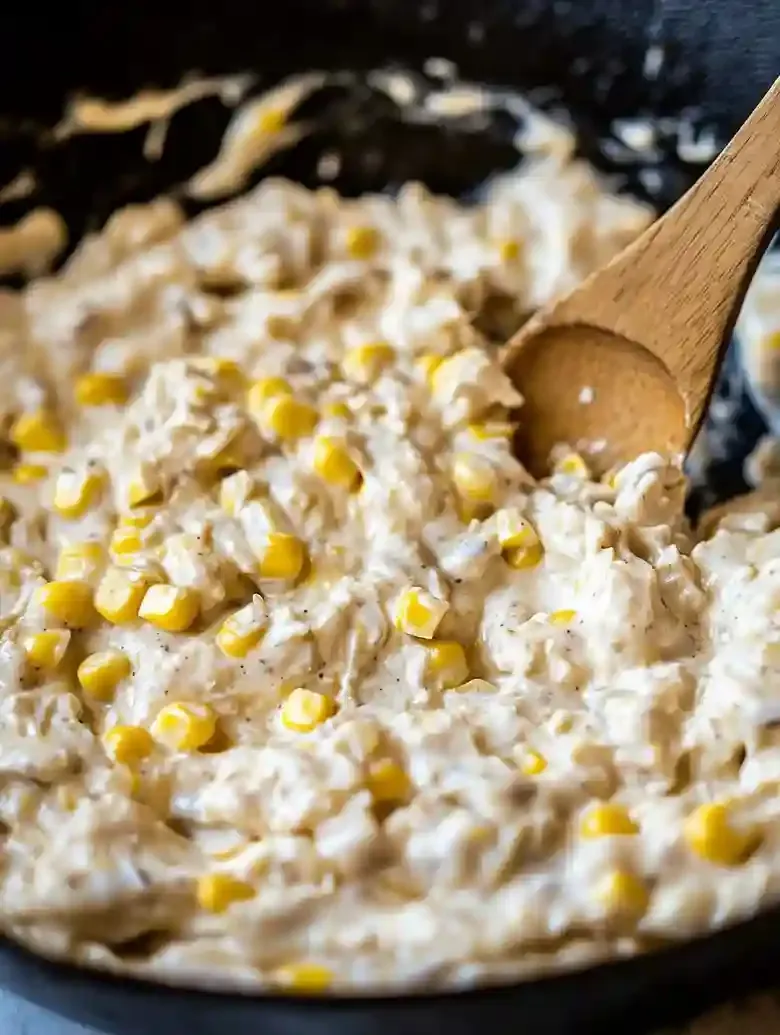Cast iron skillet with creamy sweet corn mixture and wooden spoon.