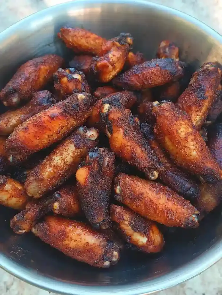 Large metal bowl with dark, seasoned chicken wings on marble surface.