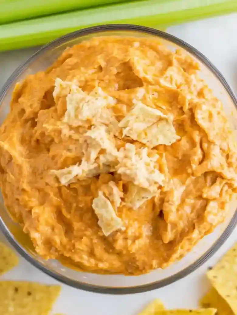 Glass bowl of orange, chunky dip garnished with shredded ingredients, chips, and celery.