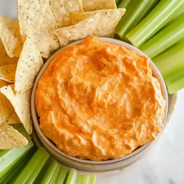 Bowl of orange dip with tortilla chips and celery sticks on marble surface.