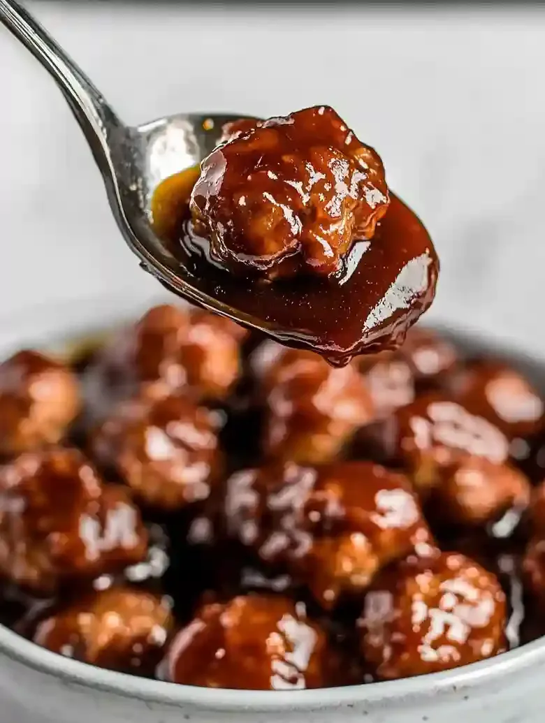 Spoonful of golden meatballs in tangy sauce lifted above a dish of meatballs.